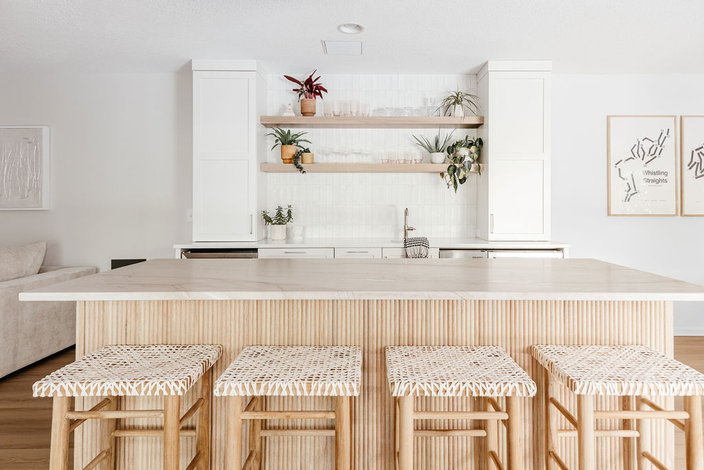 Bright, modern kitchen with a large light-stone island featuring a fluted wood base, four woven bar stools, white cabinetry, open wood shelves with plants and glassware, and a clean white tile backsplash.