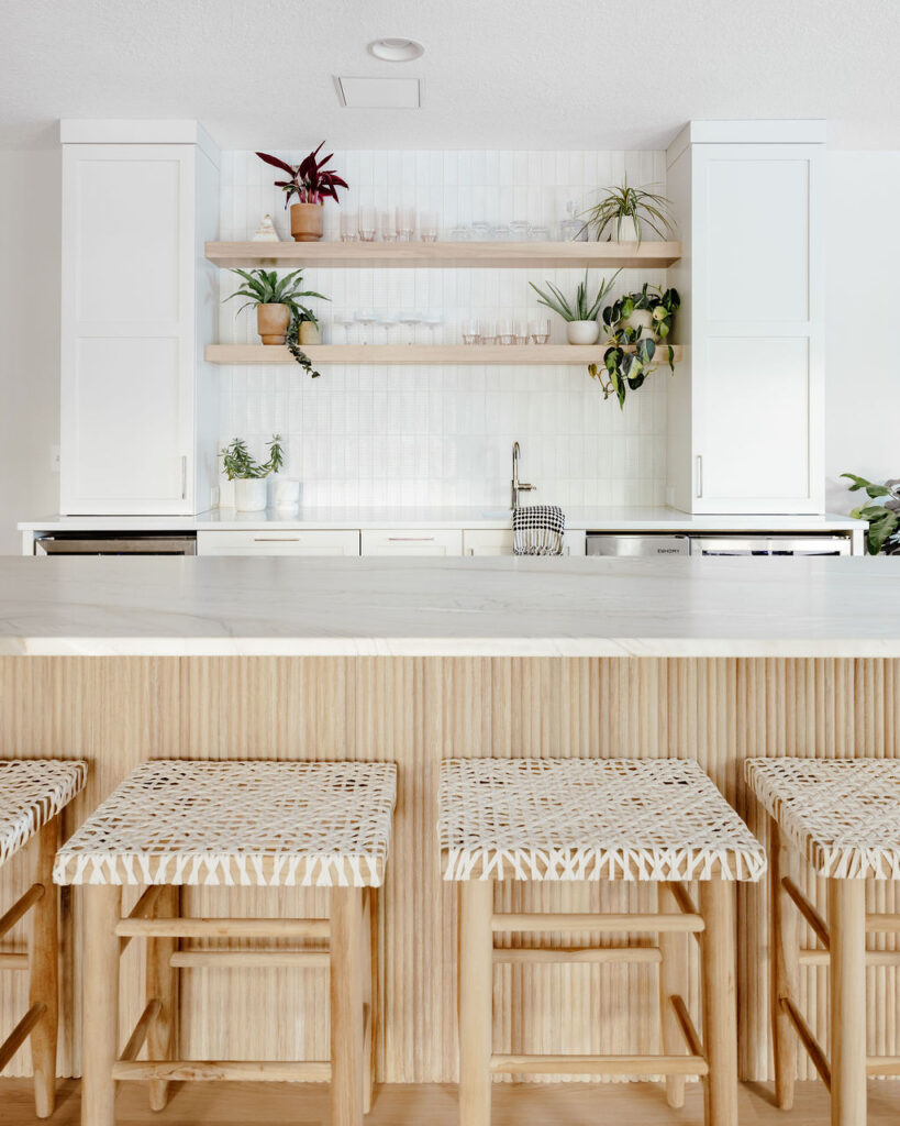 Minimalist kitchen island with a light stone countertop and vertical fluted wood base, paired with woven natural-wood bar stools; white cabinetry, open wood shelves with plants and glassware, and a white tiled backsplash in the background.