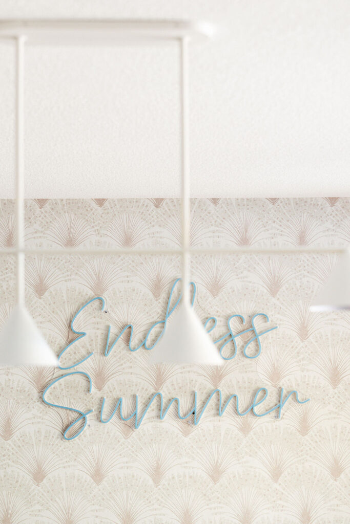 Close-up of a wallpapered accent wall with a light art deco fan motif, featuring a blue neon sign reading “Endless Summer,” partially framed by white pendant lights in the foreground.