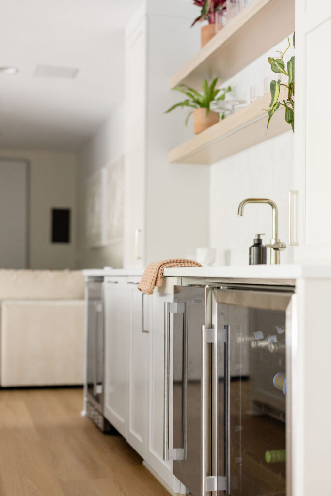Low-angle view of a bright, modern kitchenette with white cabinetry, open wood shelves holding plants and glassware, a sleek faucet and sink, and an under-counter glass-front beverage fridge, opening toward a cozy living area in the background.