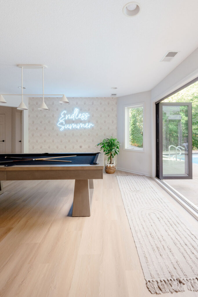 Bright, airy game room with a modern pool table on light wood floors, a minimalist white pendant light, soft patterned wallpaper with a glowing “Endless Summer” neon sign, a potted plant, and large sliding glass doors opening to a sunny pool patio.