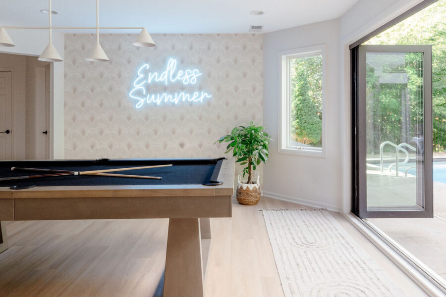 Bright, airy game room with a modern pool table on light wood floors, a minimalist white pendant light, soft patterned wallpaper with a glowing “Endless Summer” neon sign, a potted plant, and large sliding glass doors opening to a sunny pool patio.