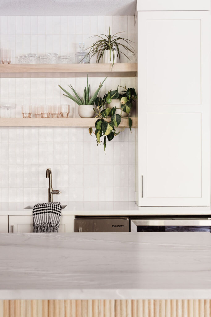 Bright, minimalist kitchen with white cabinets and a tiled backsplash, featuring two open wooden shelves holding glassware and potted plants, a small sink with a towel, and a light stone countertop in the foreground.
