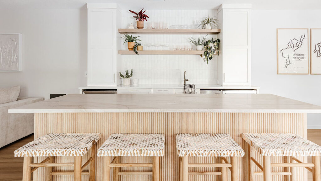 Light, modern kitchen featuring a marble island with vertical wood paneling, woven counter stools, white cabinets, and open wood shelves styled with plants and glassware.