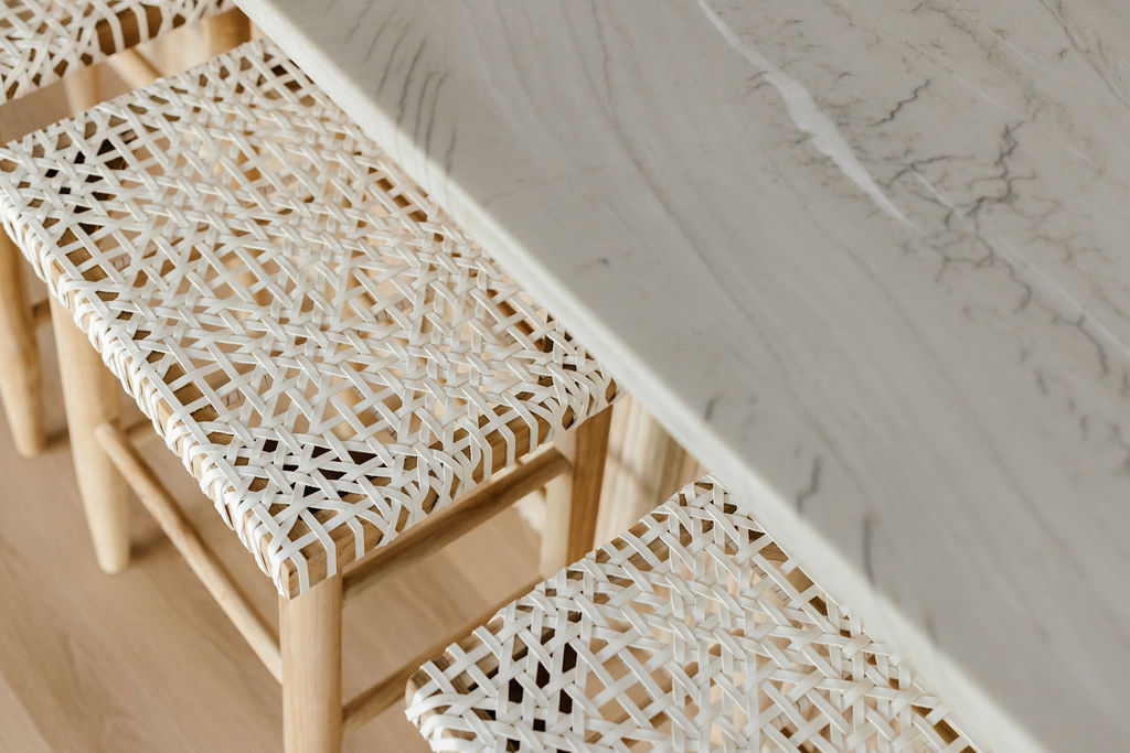 Close-up of woven white counter stools with light wood frames beneath a marble kitchen island and light wood flooring.