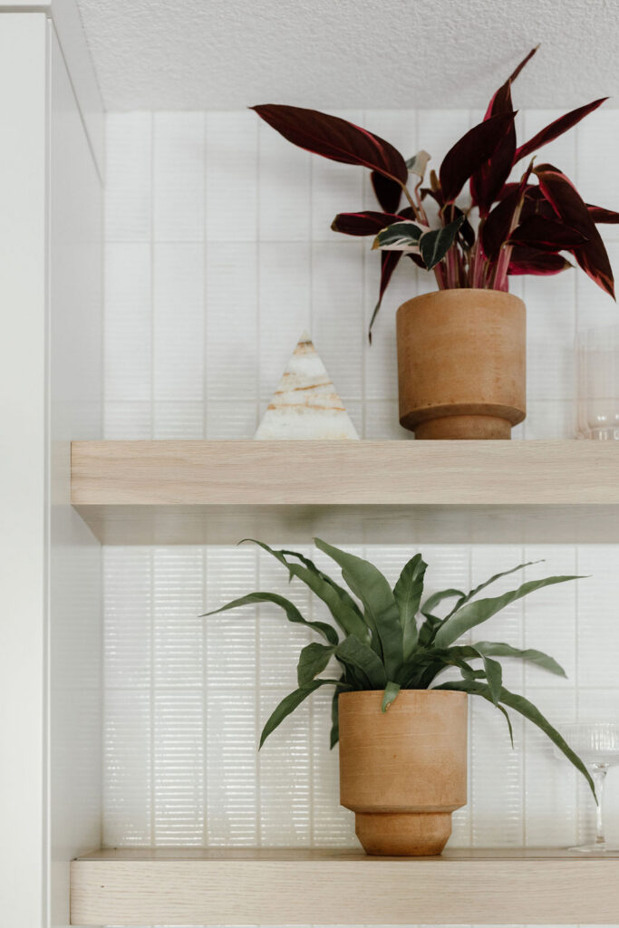 Two light wood floating shelves against a white vertical tile backsplash holding terracotta pots with plants and a small decorative stone pyramid on the upper shelf.