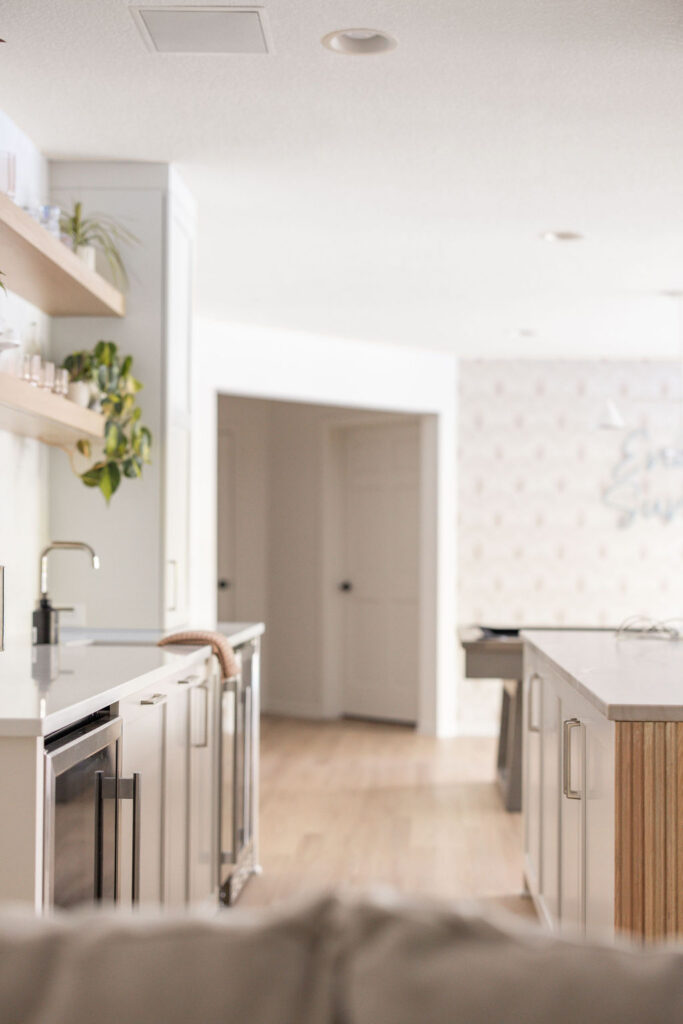 Bright modern kitchen interior with a central island, light wood floors, white cabinetry, open shelving with plants, and a doorway leading to a hallway in the background.