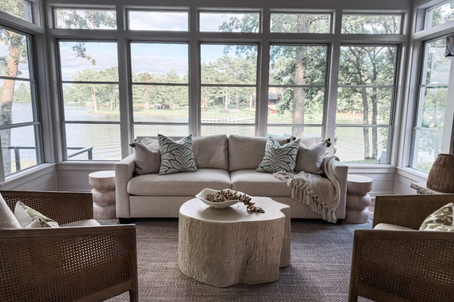 Sunroom over looking the lake with a cream couch, palm throw pillows and two rattan accent chairs.