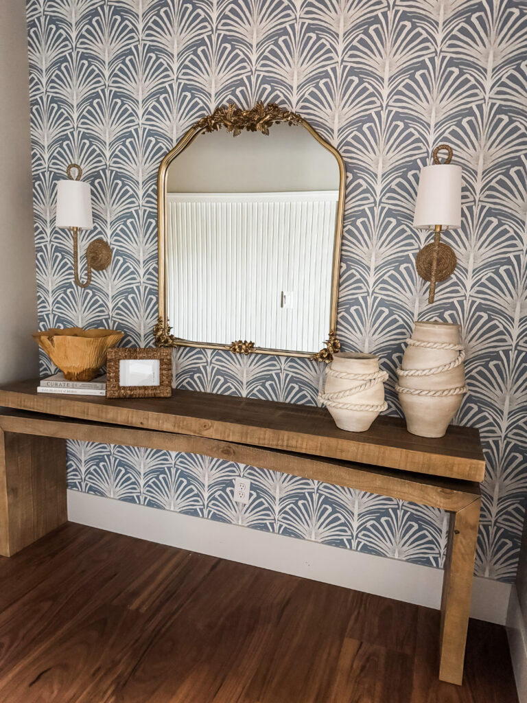 A gold mirror anchored with two gold sconces on blue printed wallpaper with a wooden console table beneath.
