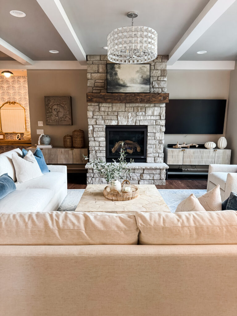 Stylish living room interior with a stone fireplace centered on the back wall, flanked by built-in shelving and a TV. A modern chandelier hangs from the ceiling, and a light neutral sectional sofa and wood coffee table are arranged in front of the fireplace. Decorative accents include a framed painting above the mantel, throw pillows, and small tabletop decor.