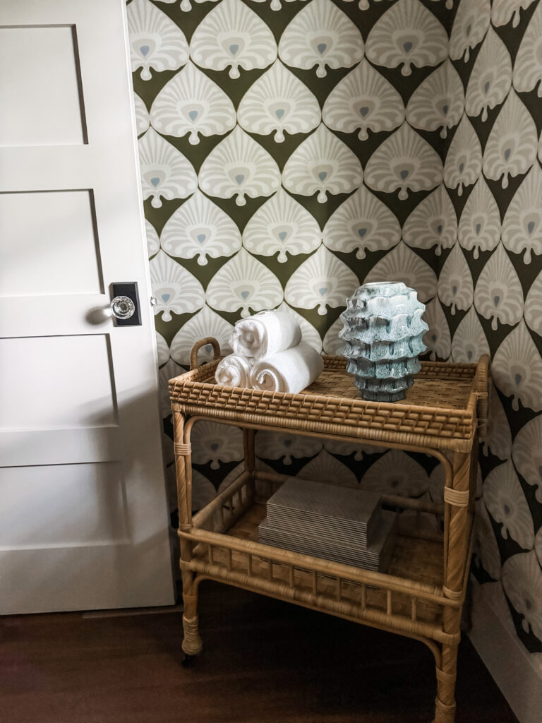 Small rattan side table against patterned green and white wallpaper holding three rolled white towels and a textured blue-green ceramic vase, next to a white paneled door.