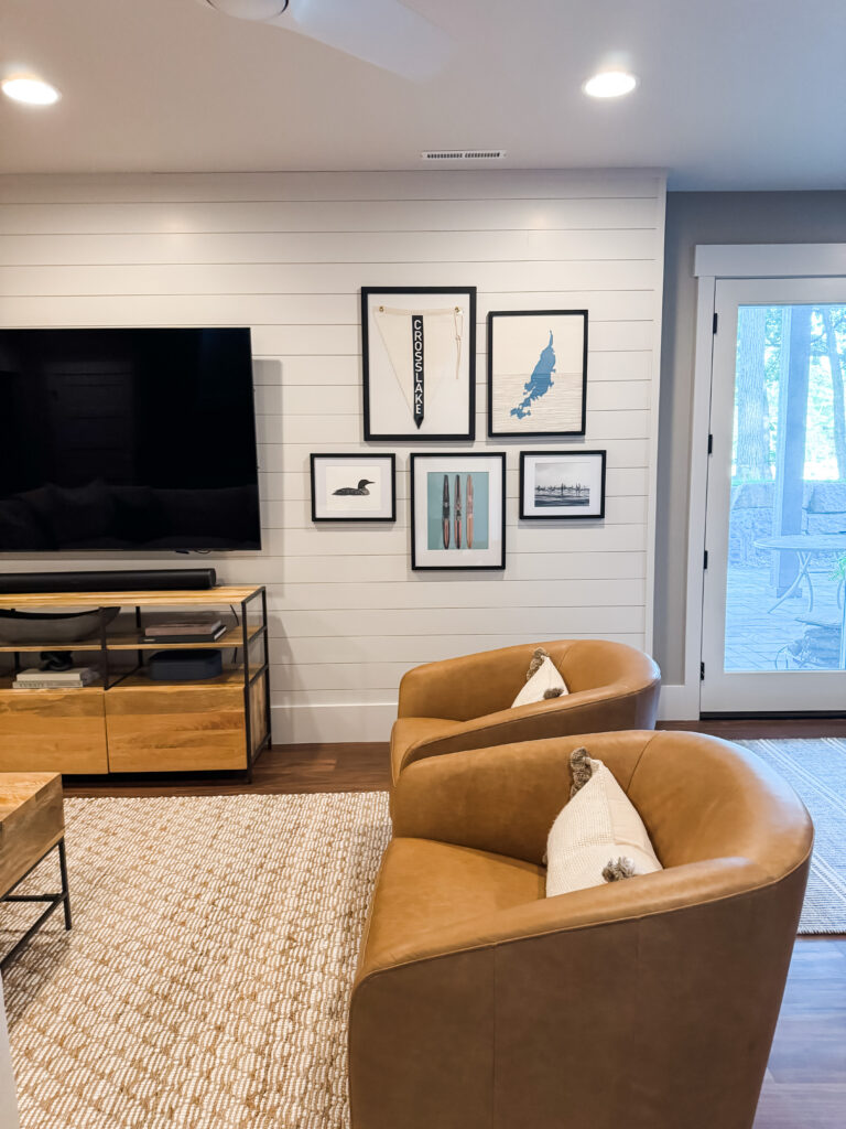Bright living room with two tan leather swivel chairs with white pillows on a woven rug, a TV mounted above a wooden media console to the left, and large glass doors to the outside on the right; a gallery wall of framed artwork above white shiplap paneling.