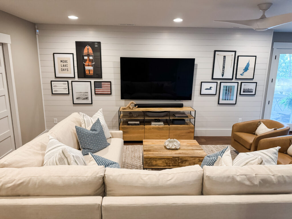 Stylish family room interior showing a large cream sectional sofa with blue and white throw pillows facing a wall-mounted flat-screen TV above a wooden media console; a wooden coffee table in front, a tan leather chair to the right, and a gallery wall of framed art on white shiplap paneling.