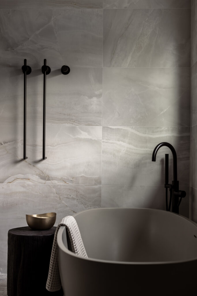 Freestanding gray bathtub with matte black wall-mounted tub filler and controls, set against large-format gray stone-look tile with soft veining; a dark wood stool with a small metallic bowl sits beside the tub, and a textured towel drapes over the edge in a moody, minimalist bathroom.
